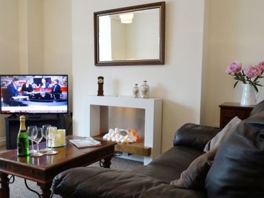 A living room with a brown leather sofa a wooden coffee table with wine glasses and a bottle of champagne a fireplace a TV showing a news program and a side table with pink flowers in a vase at 2 Carlton Mews in Exmouth