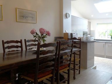 A dining area with a wooden table and chairs with cushions and a vase of flowers next to a kitchen with white cabinets and a countertop at 2 Carlton Mews in Exmouth