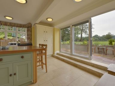 A kitchen area with wooden chairs and a green kitchen island next to large sliding glass doors opening to a patio at Abbots Manor in Combe Raleigh near Honiton