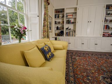 A room with a yellow sofa and cushions next to a window with flowers and built in white bookshelves filled with books and decorative items at Abbots Manor Combe Raleigh near Honiton