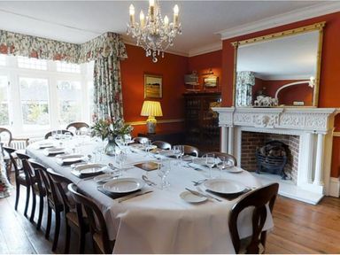 A dining room with a long table set for a meal chairs floral curtains a chandelier a fireplace and a large mirror at Abbots Manor Combe Raleigh near Honiton