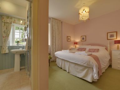 A bedroom with a double bed and two bedside tables with lamps next to an en suite bathroom with a sink and window at Abbots Manor in Combe Raleigh near Honiton