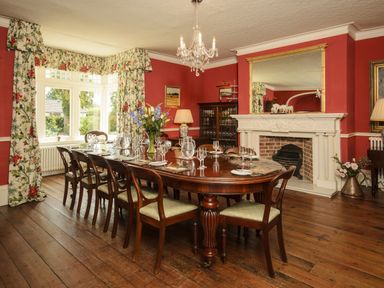 A dining room with a long wooden table set with glasses and cutlery a fireplace and floral curtains at Abbots Manor in Combe Raleigh near Honiton