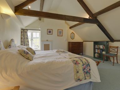 A bedroom with a double bed floral cushions and bed runners wooden beams a chest of drawers bookshelf and a chair at Abbots Manor in Combe Raleigh near Honiton