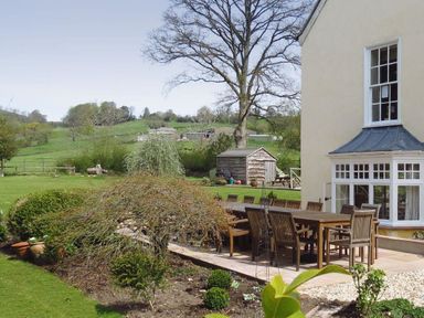 A patio with a wooden dining table and chairs next to a house and garden at Abbots Manor in Combe Raleigh near Honiton