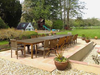 An outdoor patio with a long wooden table and chairs surrounded by plants and a lawn at Abbots Manor in Combe Raleigh near Honiton