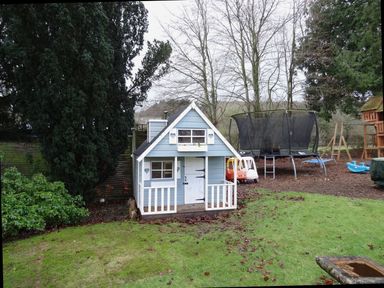 A small blue playhouse with white trim in a garden next to a trampoline and children's ride-on toys at Abbots Manor in Combe Raleigh near Honiton