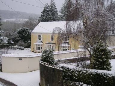A yellow house with snow on the roof and ground surrounded by trees and walls at Abbots Manor in Combe Raleigh near Honiton