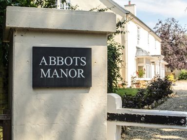 An entrance pillar with a sign reading Abbots Manor and a glimpse of a white house and garden at Abbots Manor in Combe Raleigh near Honiton
