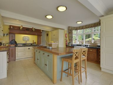 A kitchen with an island and two wooden stools near a window with chairs outside at Abbots Manor in Combe Raleigh near Honiton