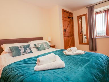 A bedroom with a double bed towels on the bed wooden door wooden framed mirror and window with vertical blinds and curtains at The Old Stable in Sidbury near Sidmouth