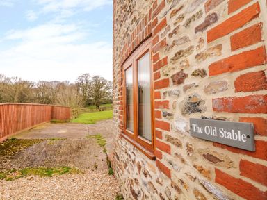 A stone and brick exterior wall with a window and a sign reading The Old Stable with a wooden fence and grassy area outside at The Old Stable in Sidbury near Sidmouth
