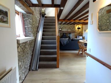 A carpeted staircase with a stair lift next to a stone wall and a living room with wooden beams and wooden chairs at Whitcombe Cottage in Honiton