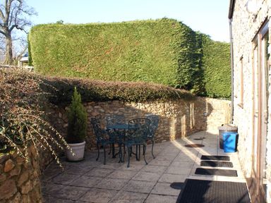 A stone patio with metal table and chairs beside a stone wall and trimmed hedge at Whitcombe Cottage in Honiton