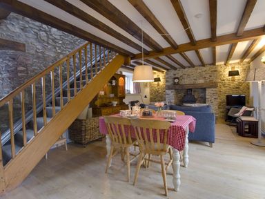 A dining table with a red checkered tablecloth and wooden chairs under a wooden beam ceiling in a room with stone walls and a fireplace at Whitcombe Cottage in Honiton