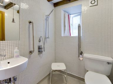 A bathroom with a wall-mounted sink a toilet a shower area with a shower stool and grab bars at Whitcombe Cottage in Honiton