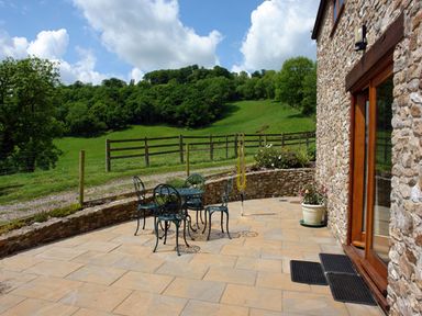 An outdoor stone patio with a metal table and four chairs next to a stone wall and a grassy hill at Swallows Cottage in Honiton