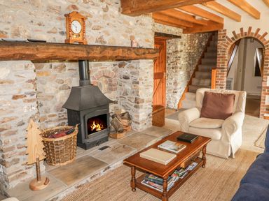 A living area with a wood stove in a stone wall fireplace a wooden coffee table with books an armchair a wicker basket and stairs at Swallows Cottage in Honiton