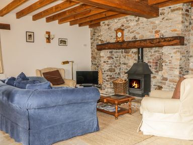 A living room with a blue sofa beige armchairs a wooden coffee table a stone fireplace with a wood stove and wooden beams on the ceiling at Swallows Cottage in Honiton