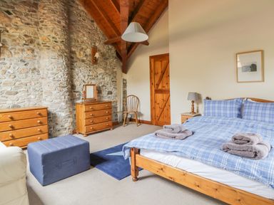 A bedroom with a wooden bed and blue checkered bedding two wooden dressers a wooden chair a blue ottoman and a stone wall at Swallows Cottage in Honiton