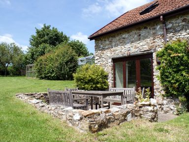 A stone house with a tiled roof and wooden doors next to a sunken patio with wooden benches and a table surrounded by a low stone wall in a grassy garden at Battens in Colyton