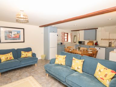 A living room with blue sofas and yellow pillows next to a kitchen with blue cabinets and a wooden dining table at Battens in Colyton