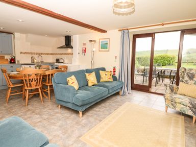 A living area with blue sofas yellow cushions a wooden dining table with chairs kitchen appliances and a glass door leading outside at Battens in Colyton
