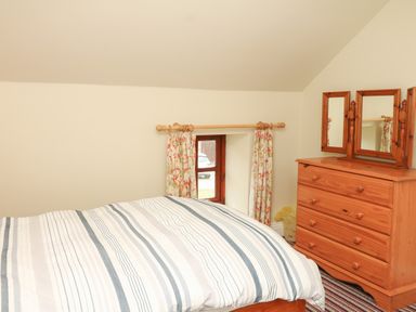 A bedroom with a bed covered by striped bedding next to a wooden dresser with a three-part mirror and floral curtains on a small window at Battens in Colyton