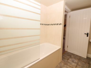 A bathroom with a bathtub that has a frosted glass shower screen and a partially open white door on tiled floor at Battens in Colyton