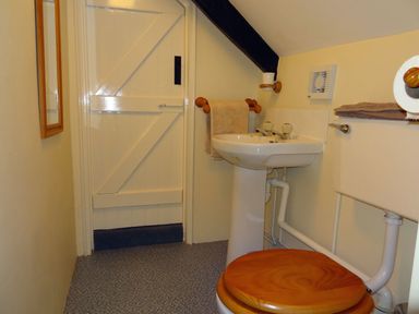 A small bathroom with a toilet featuring a wooden seat, a pedestal sink, towel holders, and a white wooden door at Lew Quarry Cottage in Lewdown