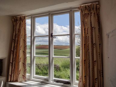 A window with patterned curtains looking out to fields and cloudy sky at West Henstill House in Sandford Devon