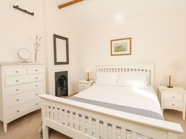 A bedroom with a white bed frame white bedside tables and lamps a white dresser with a mirror and vase a wall mirror and a framed picture at West Henstill House in Sandford Devon