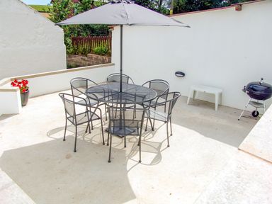 An outdoor patio with a round table and six chairs under an umbrella a white bench a barbecue and a pot with red flowers at West Henstill House in Sandford Devon