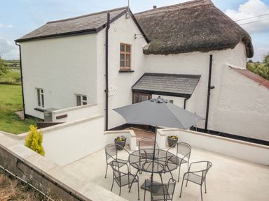 An outdoor patio with a round table and six chairs under an umbrella next to a white house with a thatched roof at West Henstill House in Sandford Devon