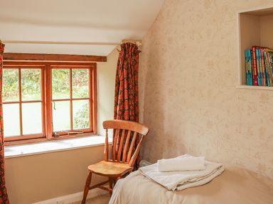 A bedroom corner with a wooden chair next to a window with red patterned curtains and a bed with folded towels and books in a wall shelf at Hope Cottage in Riddlecombe near Winkleigh
