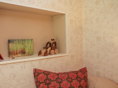 A wooden toy tractor and a photo print on a shelf above a bed with a red patterned pillow in a room with illustrated wallpaper at Hope Cottage in Riddlecombe near Winkleigh