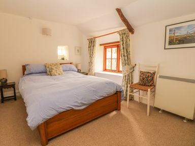 A bedroom with a wooden bed and blue bedding a window with curtains a chair with a cushion and a wall-mounted heater at Hope Cottage Riddlecombe near Winkleigh
