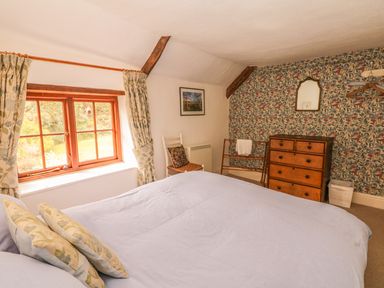 A bedroom with a bed showing pillows a wooden window with curtains a chair a chest of drawers and floral wallpaper at Hope Cottage in Riddlecombe near Winkleigh
