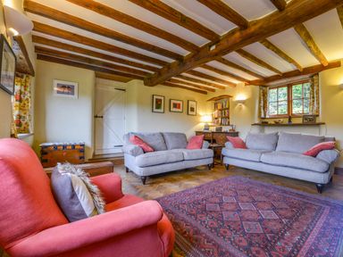 A living room with wooden ceiling beams two gray sofas a red armchair a patterned rug a wooden chest a desk with books and lamps at Hope Cottage in Riddlecombe near Winkleigh