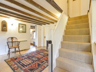 A hallway with carpeted stairs a wooden chair a patterned rug and exposed wooden ceiling beams at Hope Cottage in Riddlecombe near Winkleigh