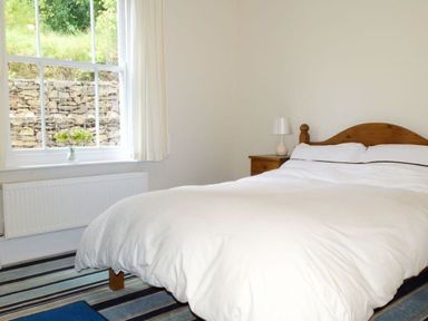 A bedroom with a wooden bed frame a white bedding a nightstand with a lamp window with curtains and a vase of flowers on the sill at Hedgelea in North Tawton