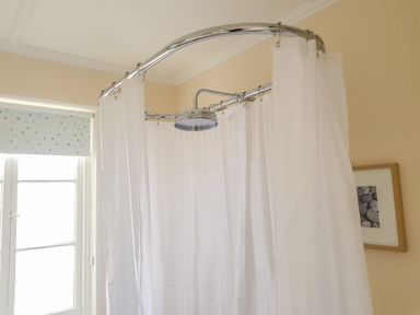 A bathroom with a ceiling-mounted round showerhead and white shower curtain near a window with a polka dot blind at Hedgelea in North Tawton