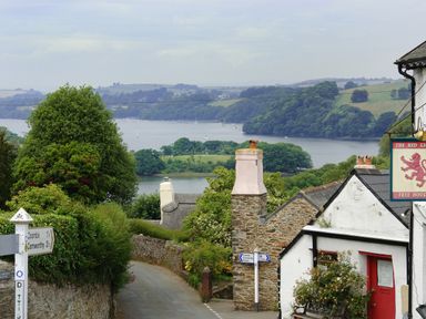 A narrow road with stone walls leading down to a river with boats surrounded by trees and hills at Linney Cottage in Dittisham