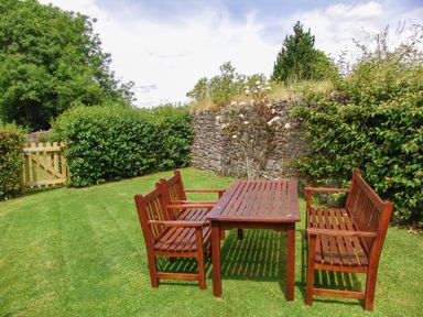 A wooden table with two benches and two chairs on grass near a stone wall and greenery at Linney Cottage in Dittisham