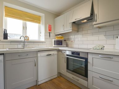 A kitchen with white cabinets a dishwasher an oven a microwave and a sink under a window at Linney Cottage in Dittisham