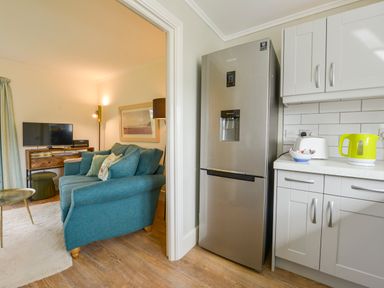 A kitchen area with a stainless steel fridge and white cabinets next to a living room with a blue sofa and a wooden TV stand at Linney Cottage in Dittisham