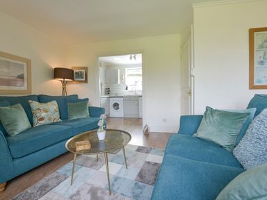 A living room with blue sofas a round brass coffee table and a view into a white kitchen at Linney Cottage in Dittisham