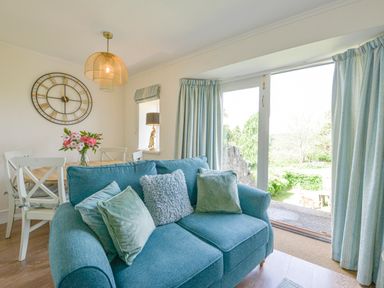 A living room with a blue sofa and cushions a dining table with chairs and flowers a large wall clock and glass doors with curtains at Linney Cottage in Dittisham