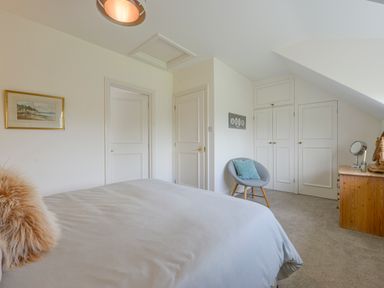 A bedroom with a bed a chair with a cushion a wooden dresser and built in wardrobes at Linney Cottage in Dittisham