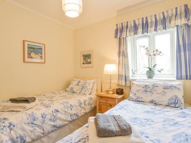 A bedroom with two single beds a wooden nightstand with a lamp between them and blue patterned curtains at Linney Cottage in Dittisham